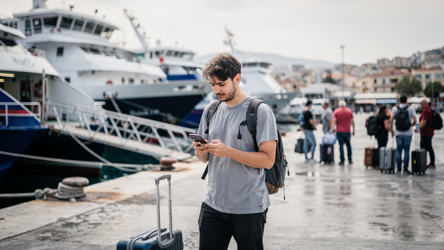A traveler uses their phone to check ferry schedules at the busy port of Piraeus, Greece, with large ferries docked in the background and other passengers waiting.