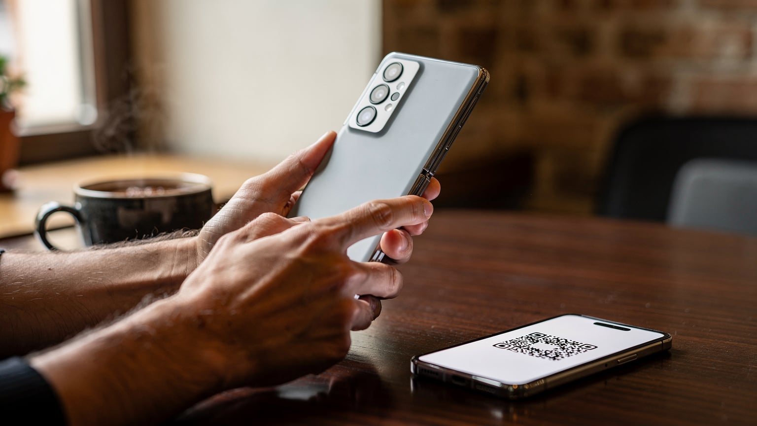 Dłonie rodzica skanują kod QR eSIM wyświetlony na ekranie drugiego smartfona, z otwartym Samsungiem Galaxy Z Fold 6 leżącym obok na drewnianym stoliku w kawiarni w Krakowie.