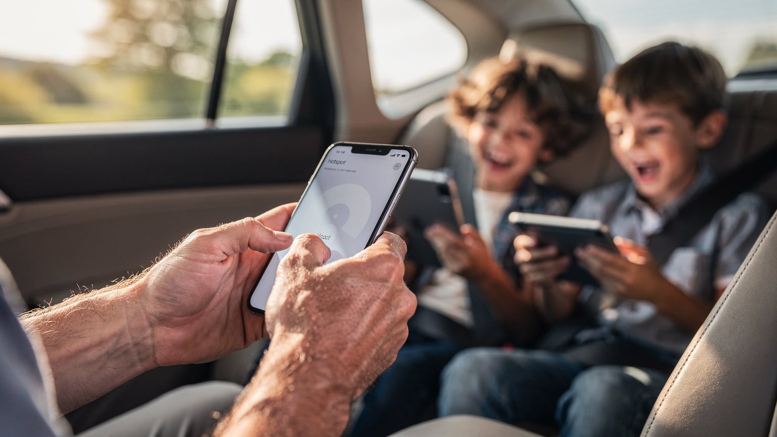 Primer plano de las manos de un padre configurando la función de hotspot en su teléfono móvil, con la interfaz de usuario en pantalla, mientras en el fondo, desenfocado, sus dos hijos ríen y juegan con sus tabletas en el asiento trasero de un coche en movimiento por la carretera.