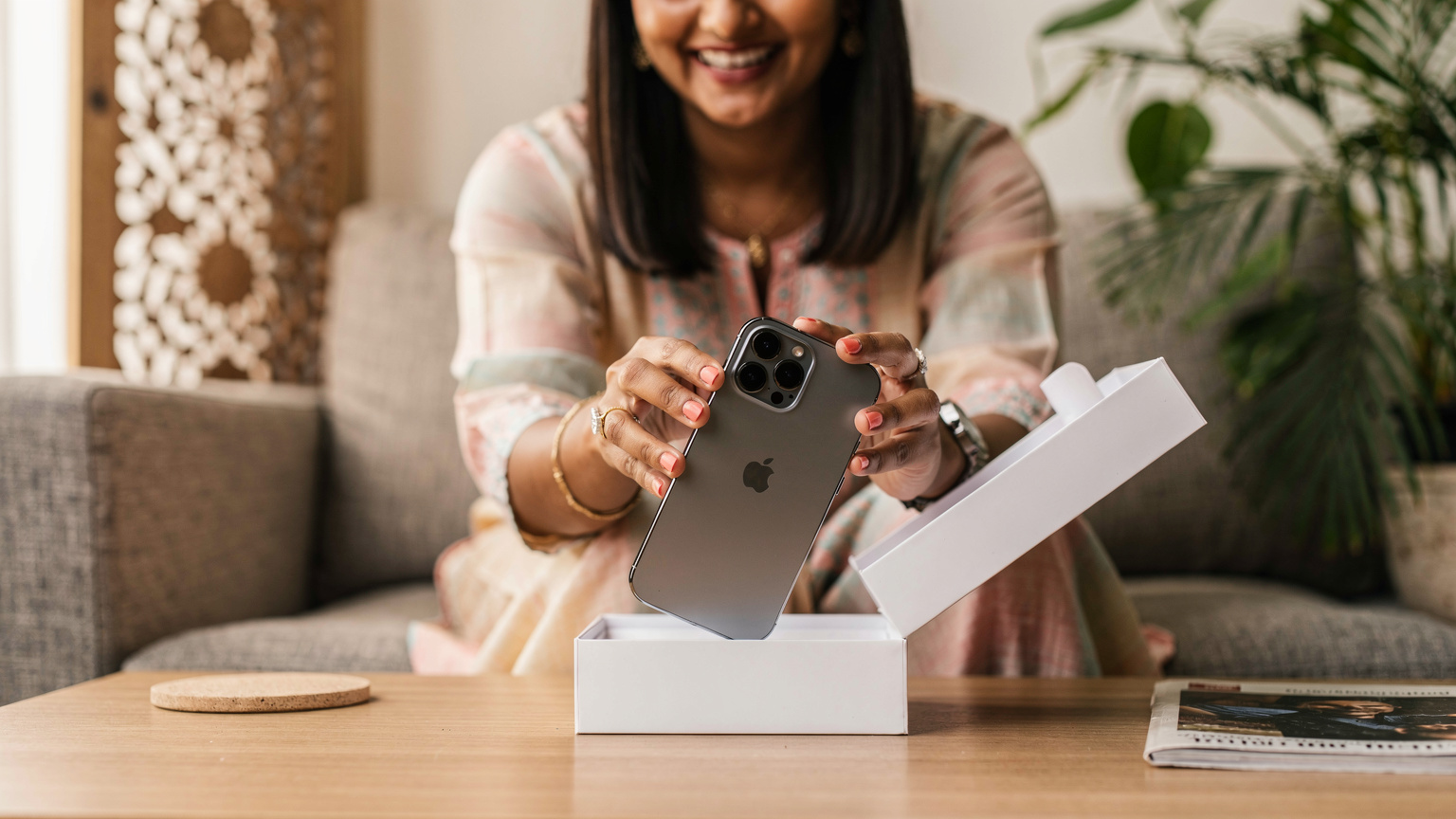 एक भारतीय महिला अपने नए iPhone 15 Pro Max को उसके बॉक्स से बाहर निकाल रही है, उसके चेहरे पर उत्सुकता और खुशी है, एक आधुनिक भारतीय घर में।