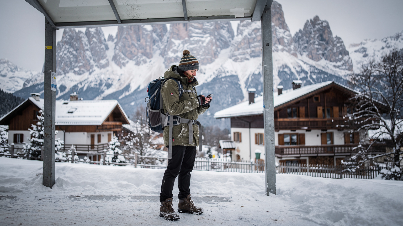 Een reiziger die bij een bushalte in Cortina d'Ampezzo op zijn telefoon een busroute controleert, met besneeuwde bergen en een typisch Italiaans dorpje op de achtergrond.