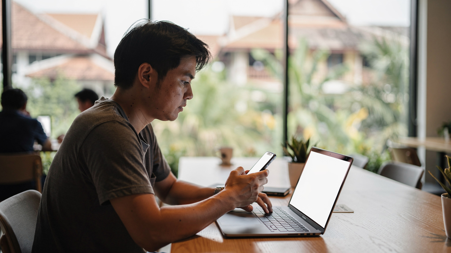 タイのチェンマイにあるコワーキングスペースで、ノートPCとスマートフォンを使いながら集中して仕事をしている日本人デジタルノマド。窓からは緑豊かな景色が見える。