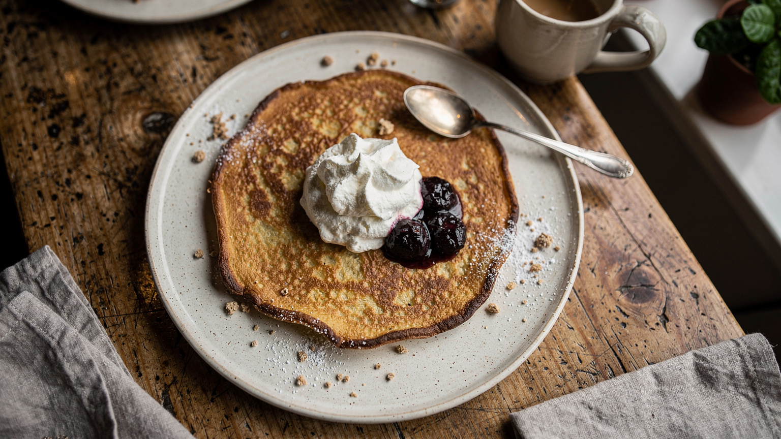 Un piatto di Ålandspannkaka, un pancake spesso, servito con marmellata di prugne e panna montata, su un tavolo di legno in una accogliente caffetteria di Mariehamn.