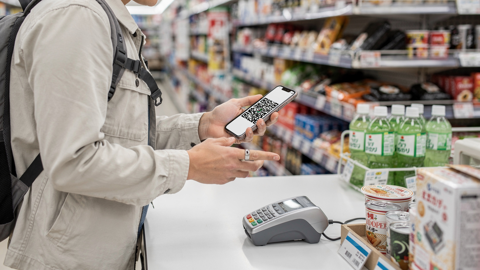 一个旅行者在东京便利店(FamilyMart)里,正在使用手机支付,手机屏幕上显示支付二维码,旁边货架上摆满了日式零食和饮料。