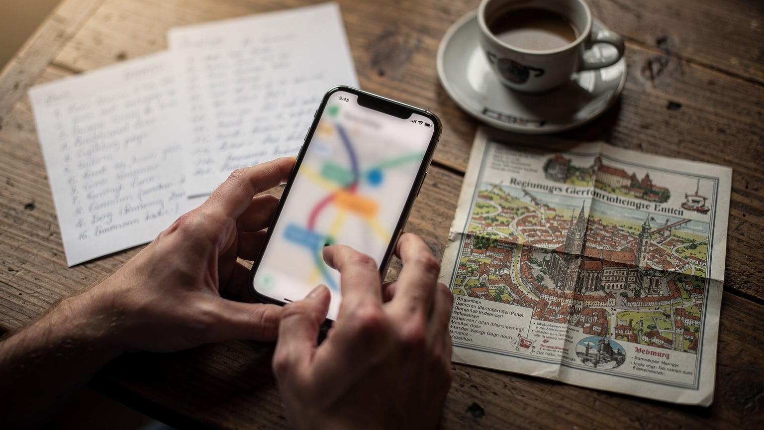 Nahaufnahme von Händen, die auf einem Smartphone eine Reiseroute im ADAC Routenplaner planen, mit Notizen und einem Stadtplan im Hintergrund.