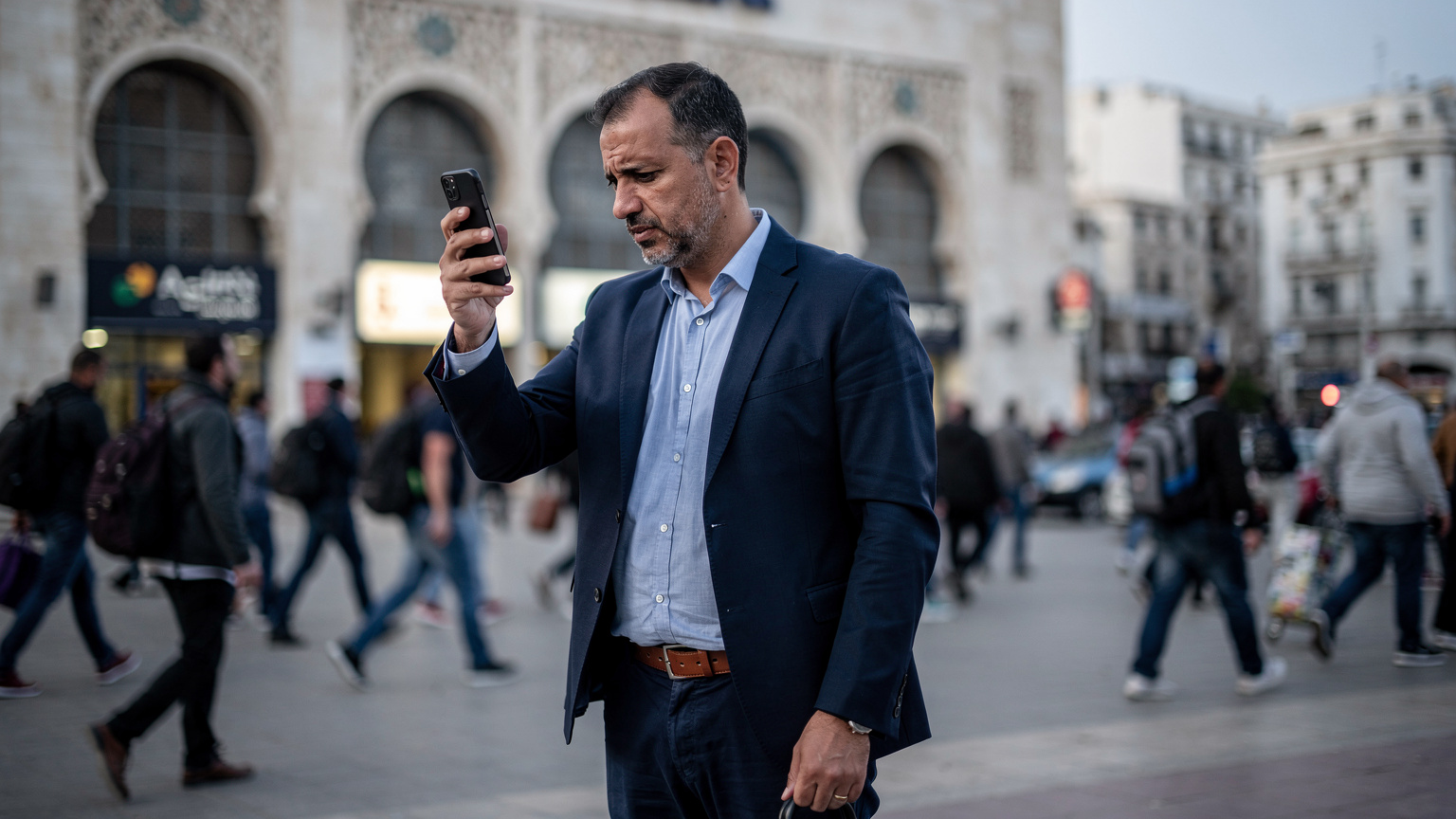 Un homme vérifie la réception de son smartphone avec un air préoccupé devant une gare ferroviaire animée à Alger, tandis que des passants se hâtent autour de lui. Le ciel est légèrement nuageux, créant une lumière douce et diffuse.