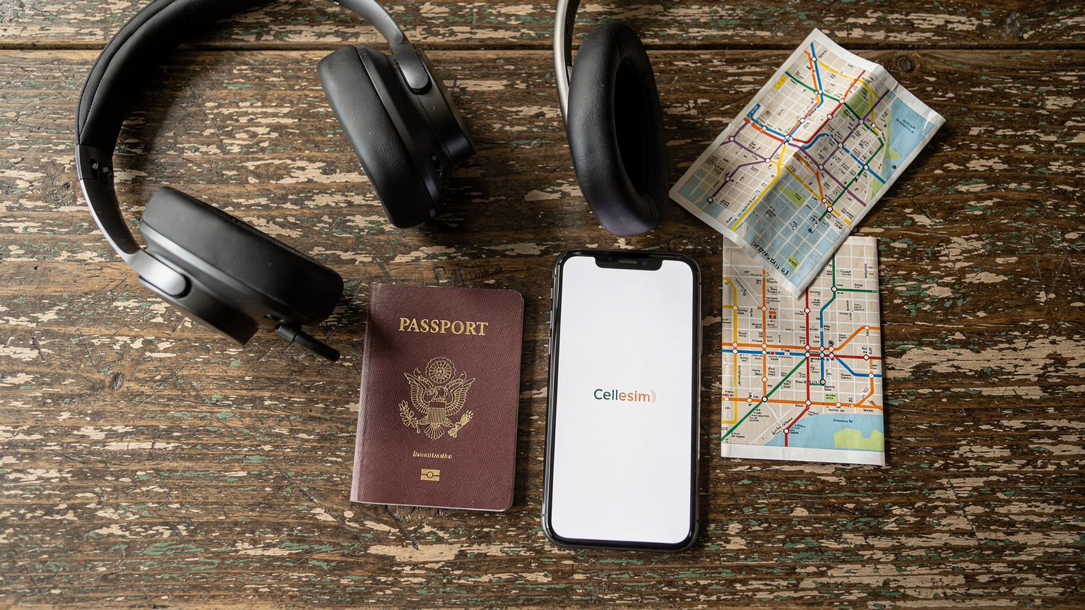 An overhead flat-lay shot of a traveler's essentials, including a passport, a smartphone showing a Cellesim logo on its screen (no readable UI), headphones, and a small, folded map, all resting on a rustic wooden table in a cafe, ready for a trip.