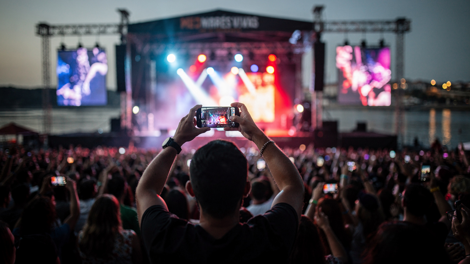 Uma pessoa de costas, segurando um smartphone na altura dos olhos, gravando um vídeo do palco principal do MEO Marés Vivas, com a multidão em segundo plano e luzes coloridas a iluminar a cena.