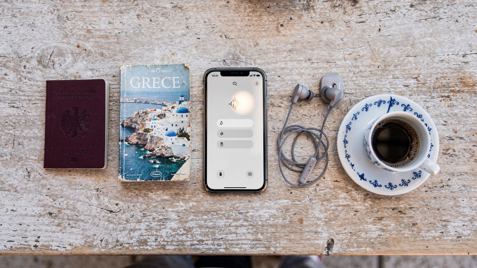 An overhead flat-lay shot of travel essentials, including a smartphone displaying network settings, a passport, a travel guide, and a Greek coffee cup on a cafe table.