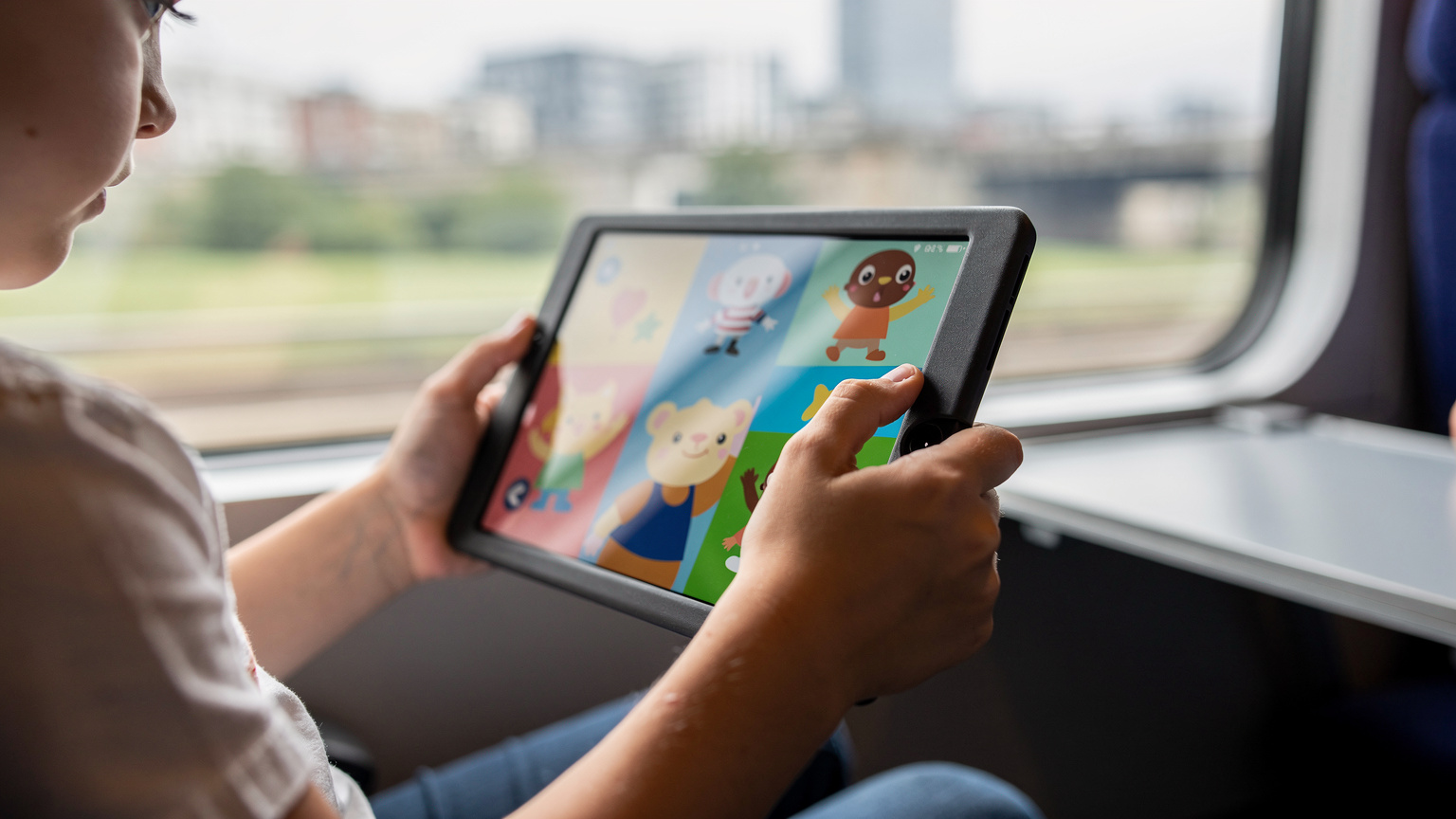 Nahaufnahme von Kinderhänden, die ein Tablet mit einer bunten, altersgerechten Offline-Spiel-App halten, während im verschwommenen Hintergrund eine Zugfahrt durch eine Stadt zu sehen ist.