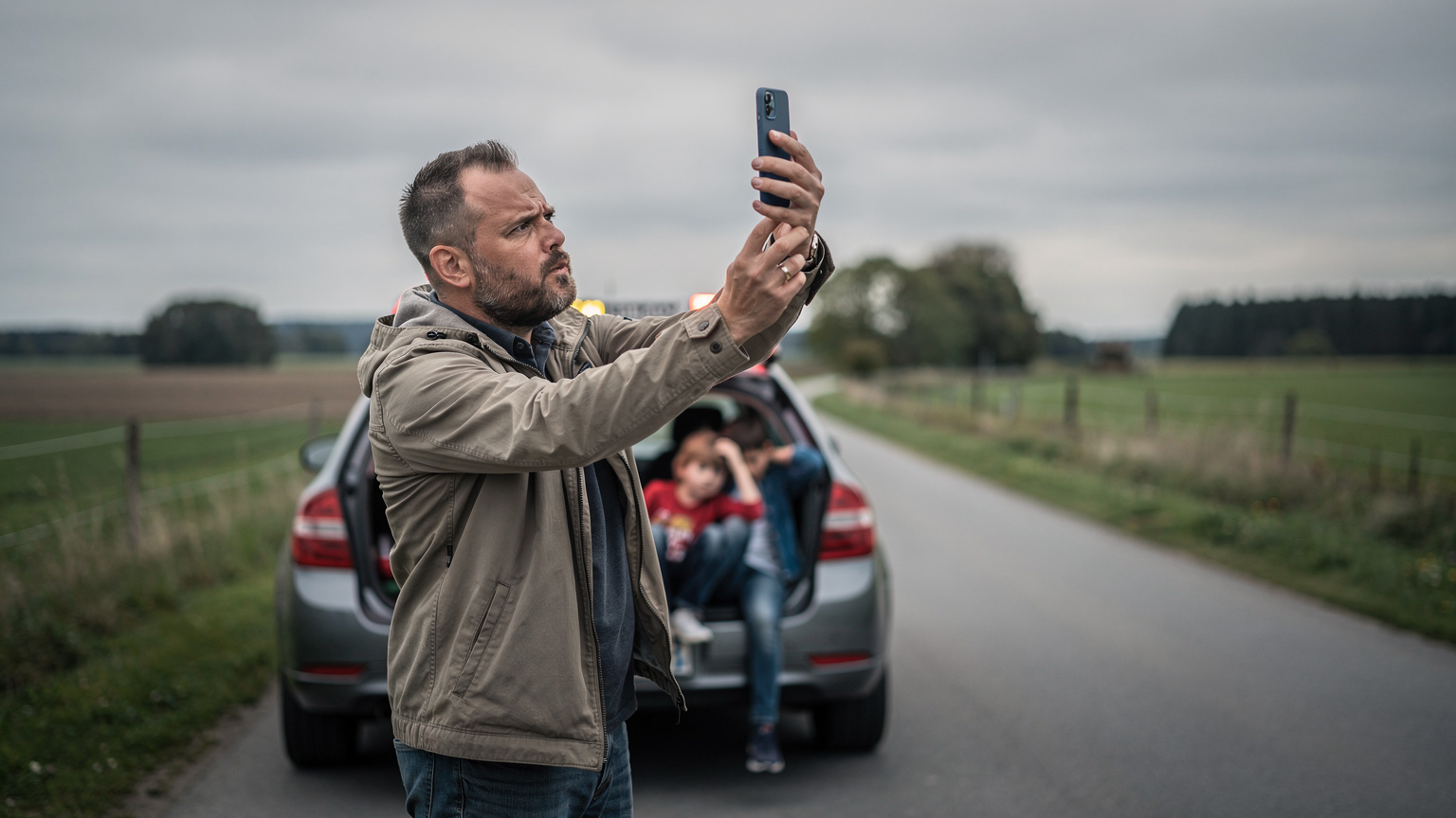 Ein verärgerter Vater versucht verzweifelt, mit seinem Smartphone am Straßenrand Empfang zu finden, während seine Kinder gelangweilt und unruhig auf der Rückbank des Autos sitzen und ihn ansehen.