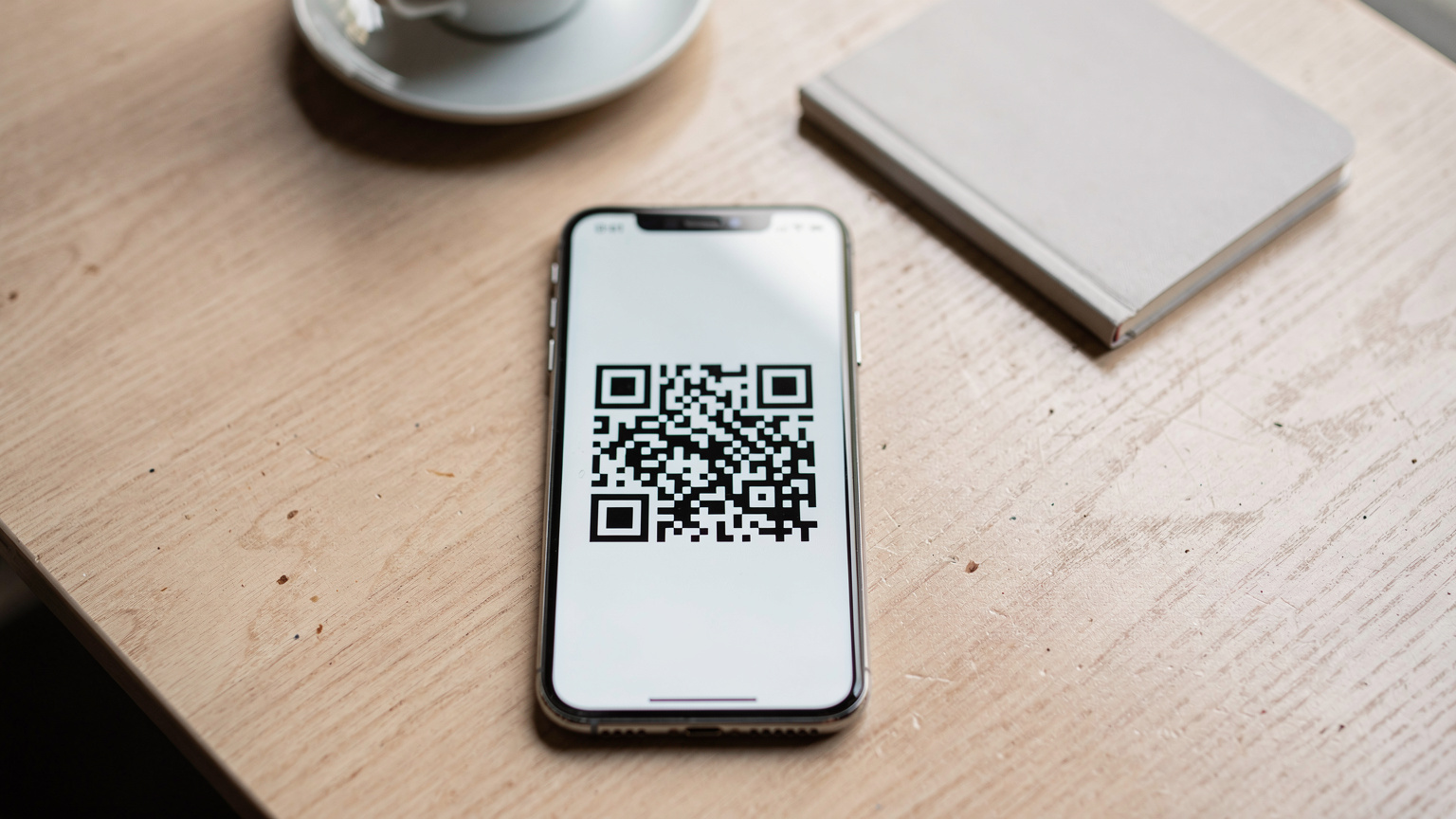 Un primo piano di un telefono che mostra un codice QR per l'attivazione di una eSIM, posato su un tavolo di legno in un caffè moderno a Lisbona, con una tazza di caffè e un taccuino sullo sfondo sfocato.