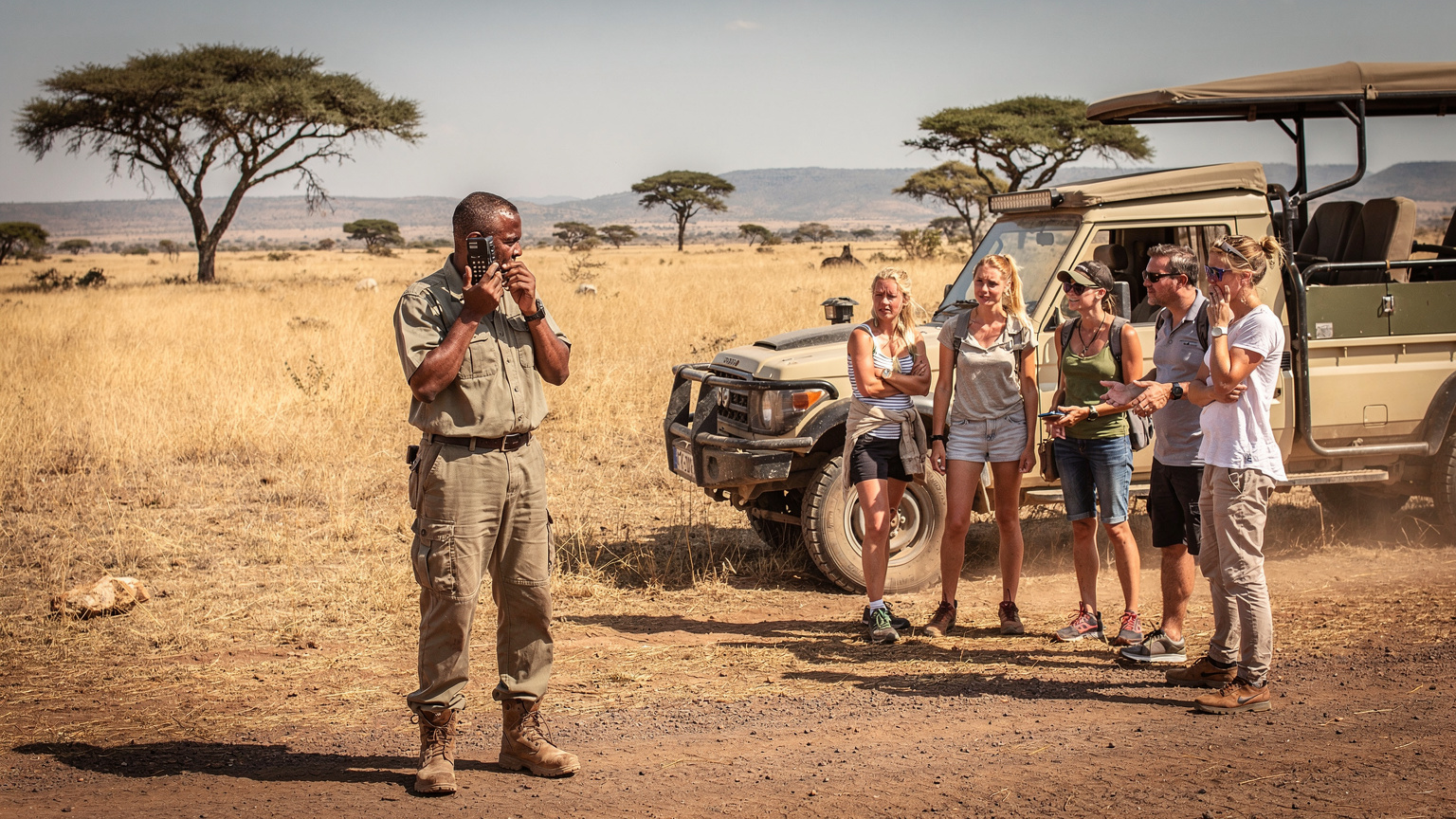En guide taler i en satellittelefon, mens en gruppe safarigæster venter ved en defekt jeep midt på en støvet grusvej i Tanzanias Serengeti nationalpark, udtryk for bekymring.