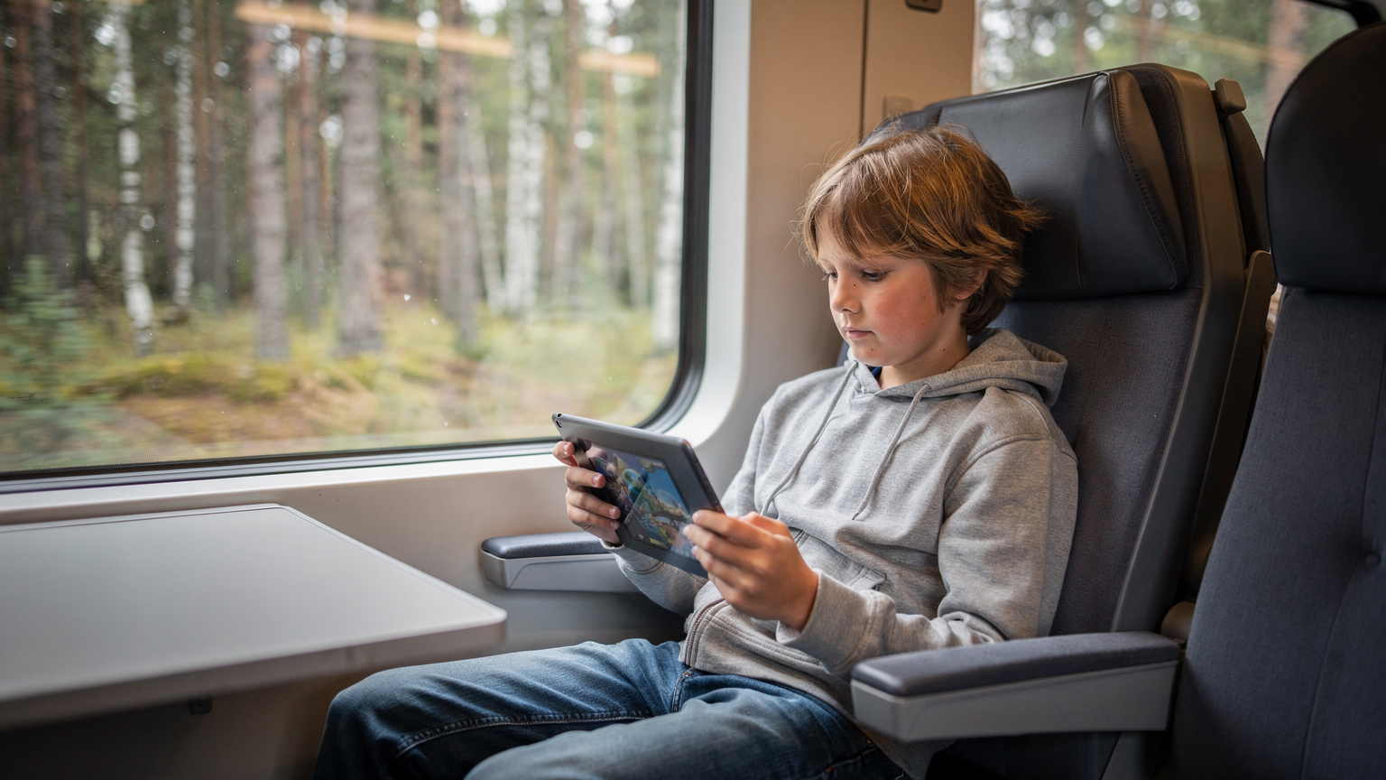Een kind, geconcentreerd bezig met een tablet waarop een offline game wordt gespeeld, zit comfortabel in een Finse trein, terwijl de bossen buiten voorbijflitsen.