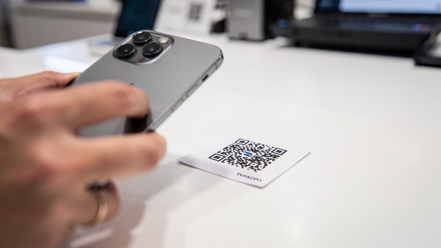 Close-up of a person's hand holding a smartphone, scanning a Turkcell eSIM QR code at a brightly lit service counter.