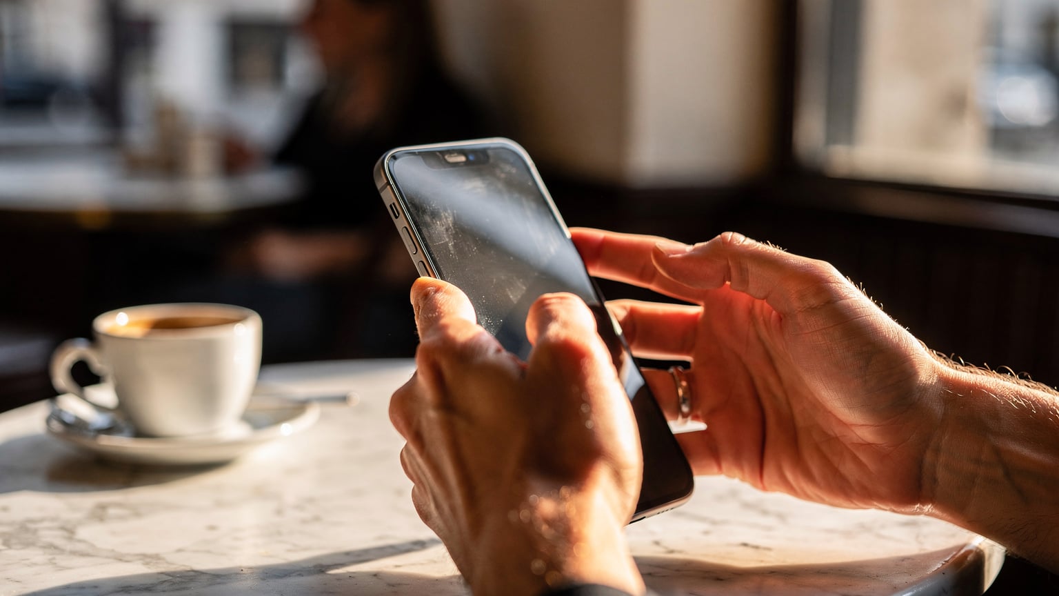 A close-up of a person's hands holding an iPhone in a sunlit Parisian cafe, dialing the *#06# code on the phone app keypad.