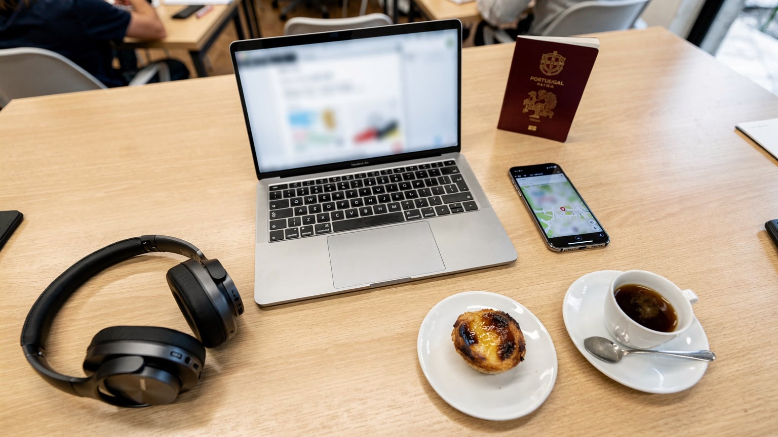 An overhead flat-lay of a traveler's desk setup in a co-working space in Porto, Portugal, featuring an open laptop, an iPhone, a passport, and a local pastry (pastel de nata) with a small cup of coffee.
