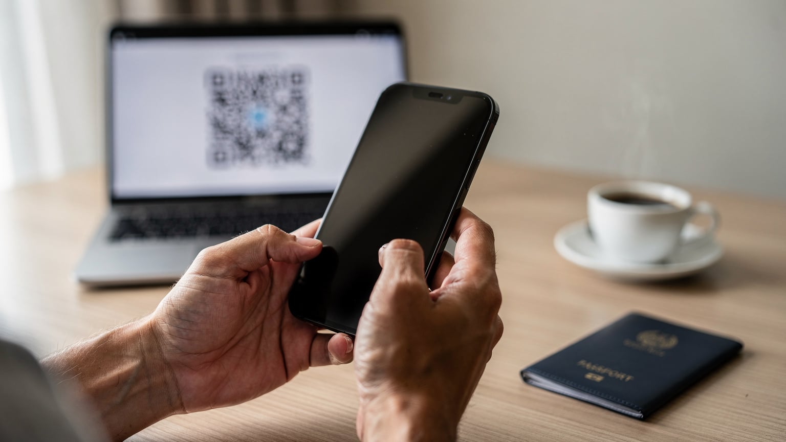 A close-up of a traveler's hands holding an iPhone, with a QR code for eSIM installation displayed on a laptop screen in the background.