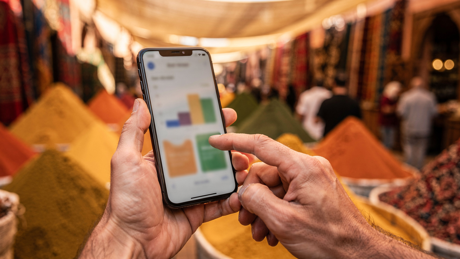Gros plan sur des mains tenant un smartphone affichant l'écran d'utilisation des données, avec l'arrière-plan flou d'un marché traditionnel marocain animé et coloré.