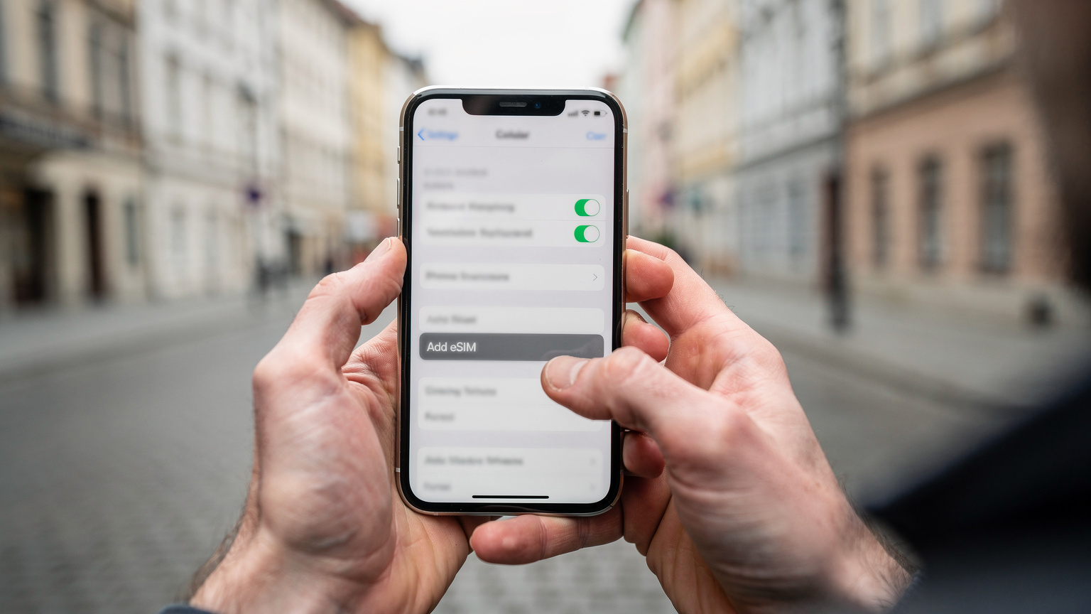 Nahaufnahme von Händen, die ein iPhone 11 halten, auf dessen Bildschirm die Mobilfunkeinstellungen mit der Option zum Hinzufügen einer eSIM angezeigt werden. Der Hintergrund ist leicht unscharf und zeigt eine europäische Altstadt.