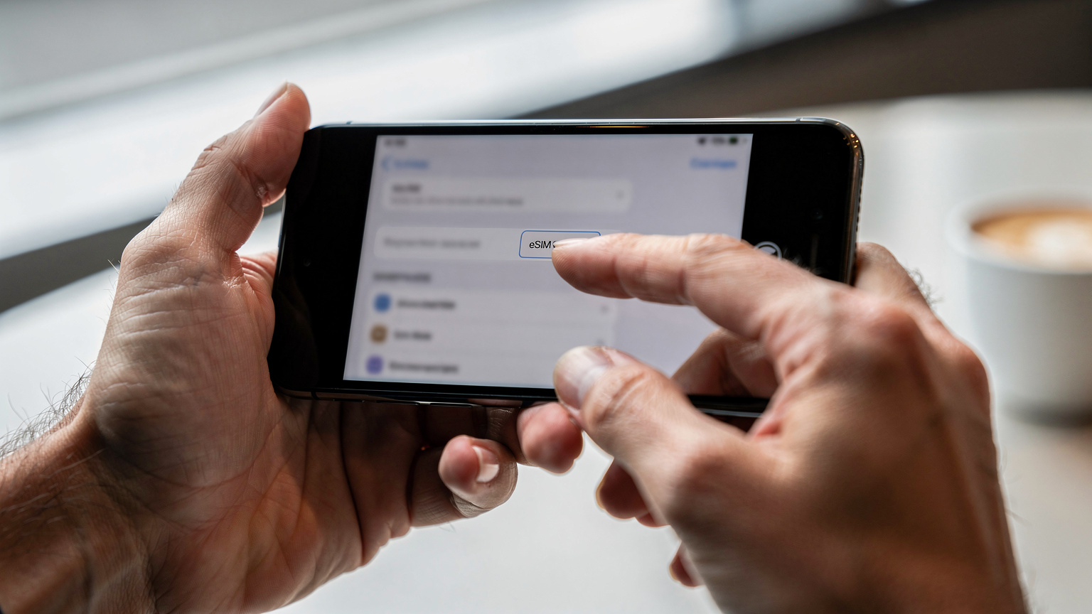 A close-up of hands holding a smartphone, displaying the phone's settings menu with an eSIM profile being added, possibly in a café or airport lounge.
