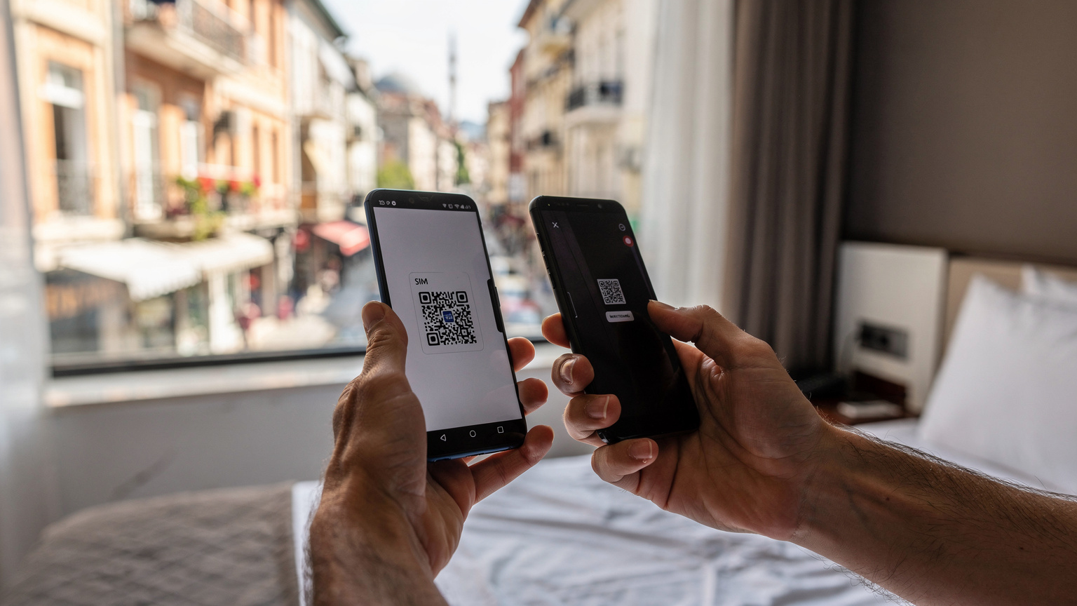 Руки тримають смартфон з QR-кодом eSIM на екрані, а інша рука використовує інший телефон для сканування цього коду, на тлі вікна готелю в районі Бейоглу, Стамбул.