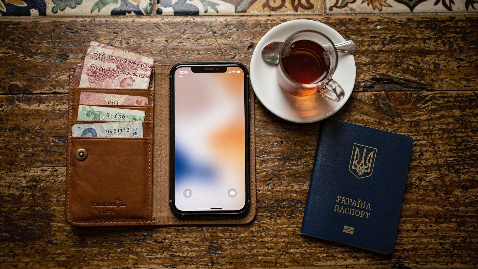 Вид зверху на стіл у кафе в районі Фатіх, Стамбул, де лежить відкритий гаманець з кількома турецькими лірами, смартфон з активним екраном, склянка турецького чаю та паспорт.