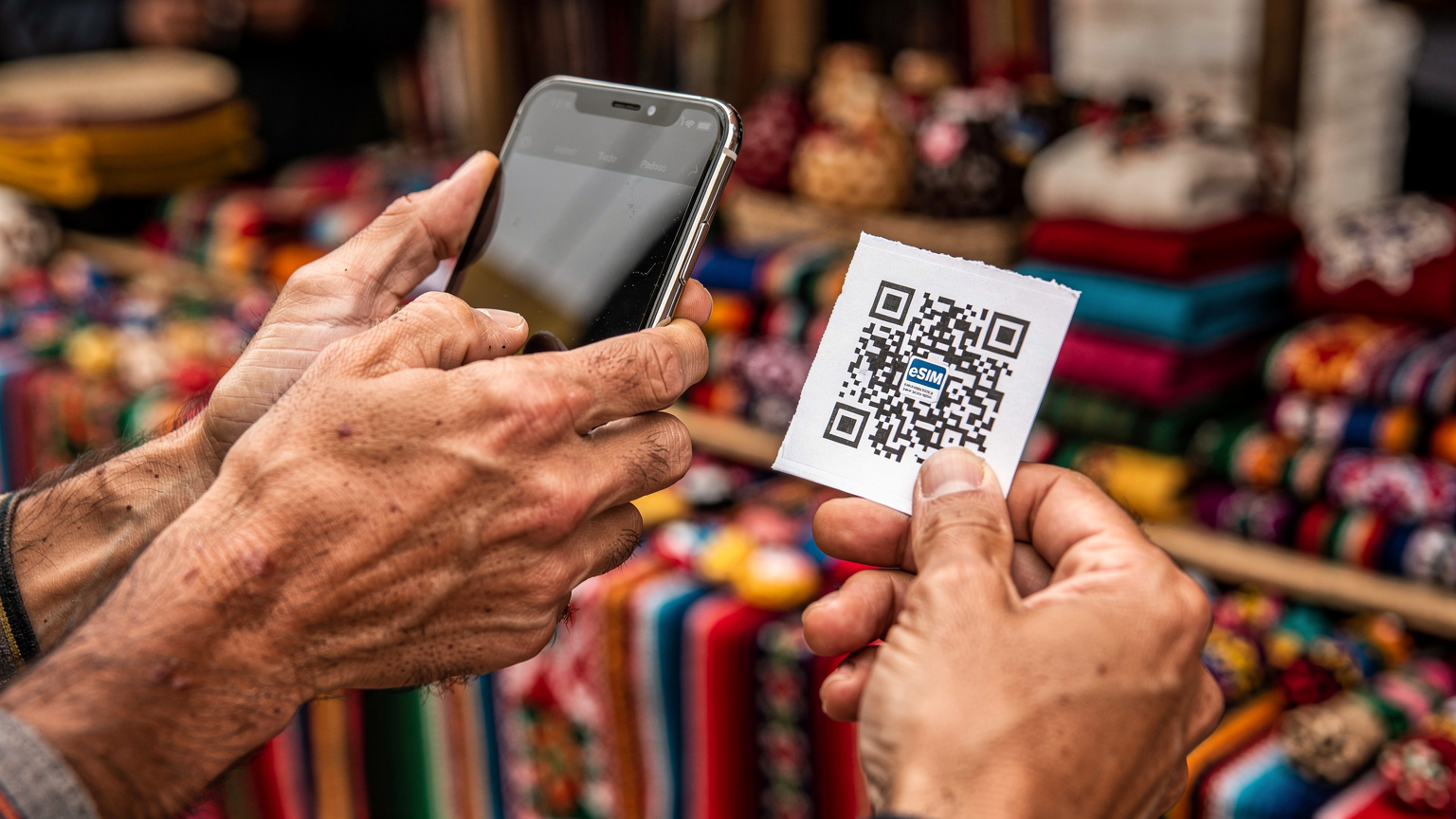 Close-up of a traveler's hands holding a smartphone, with a Cellesim QR code on a piece of paper, against a blurred backdrop of a Bolivian market stall with colorful textiles.