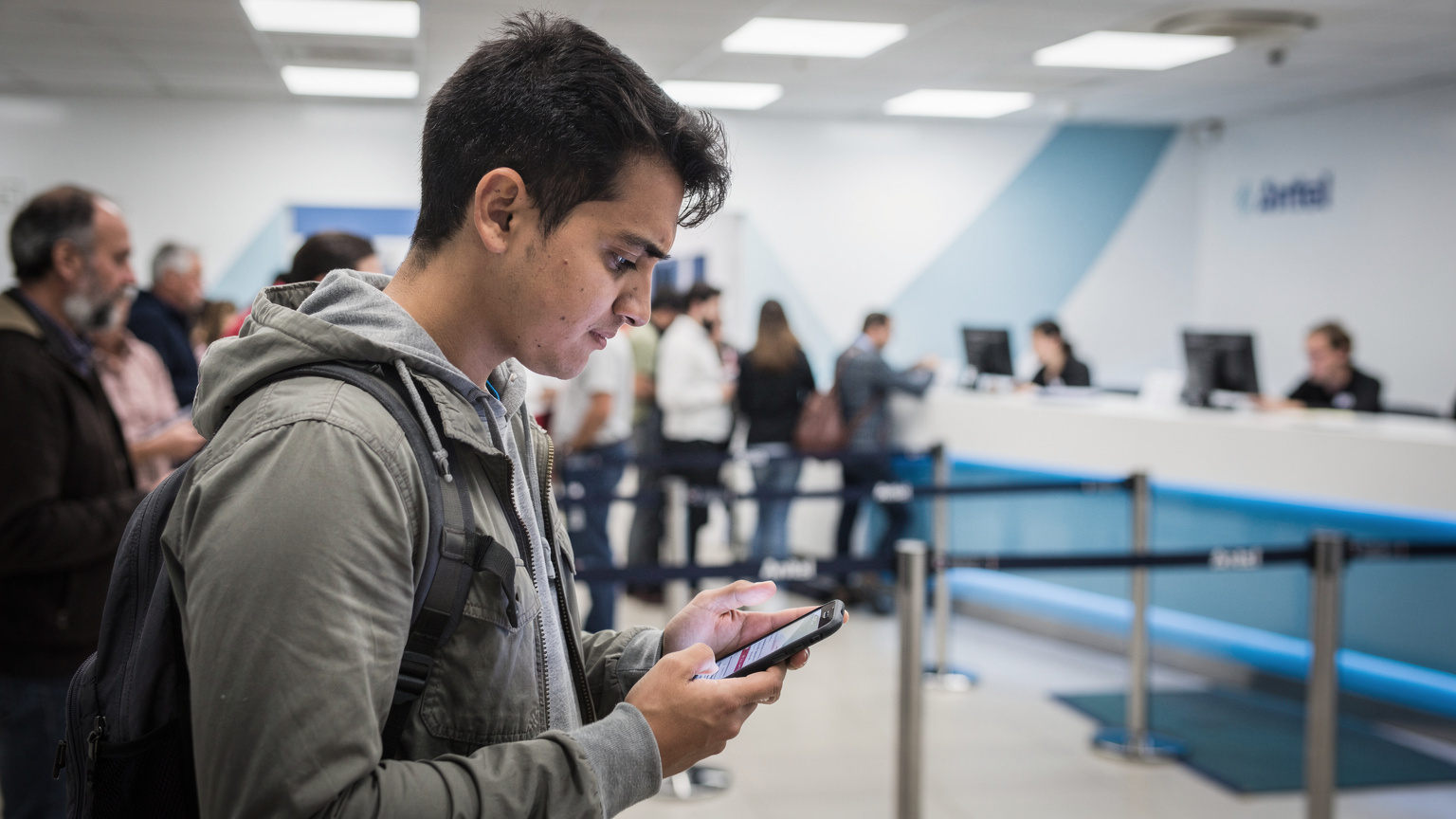 Un viajero revisa su teléfono con una expresión de frustración mientras espera en una larga fila en el centro de atención al cliente de Antel en el centro de Montevideo, con otros clientes locales al fondo.