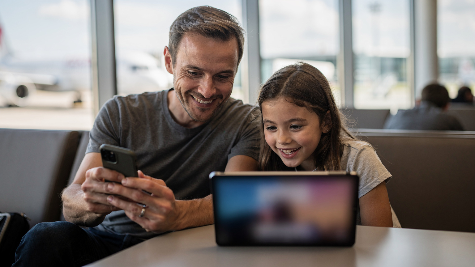 Un père et sa fille souriants préparent leurs appareils en téléchargeant des films sur une tablette avant un vol, dans un salon de départ d'aéroport moderne à Zurich. Le père a un smartphone à la main, et la fille regarde l'écran de la tablette avec anticipation.