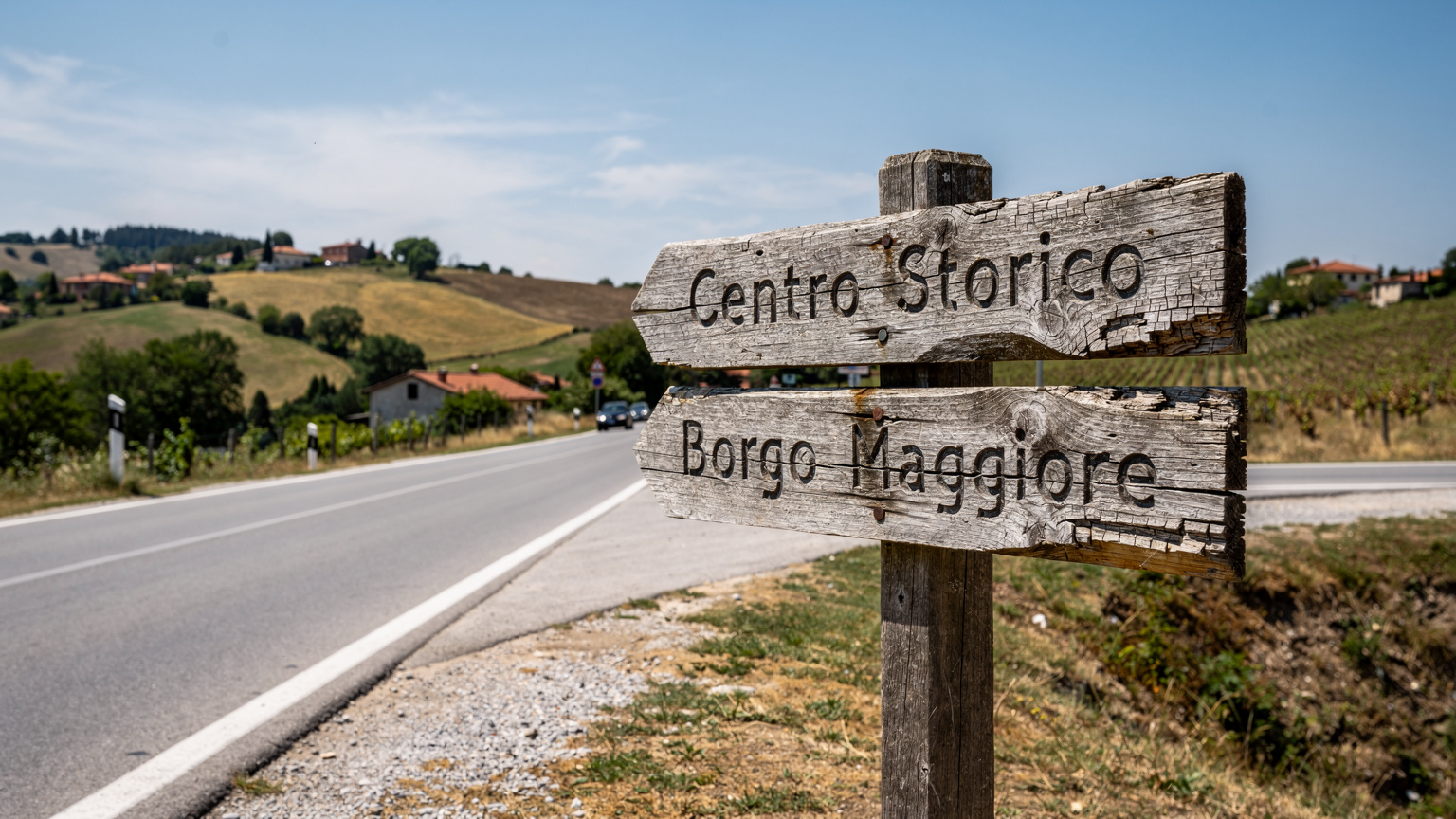 Un cartello stradale in legno indica le direzioni per il centro storico di San Marino e Borgo Maggiore, con un leggero traffico di auto in lontananza sotto un cielo azzurro e sereno.