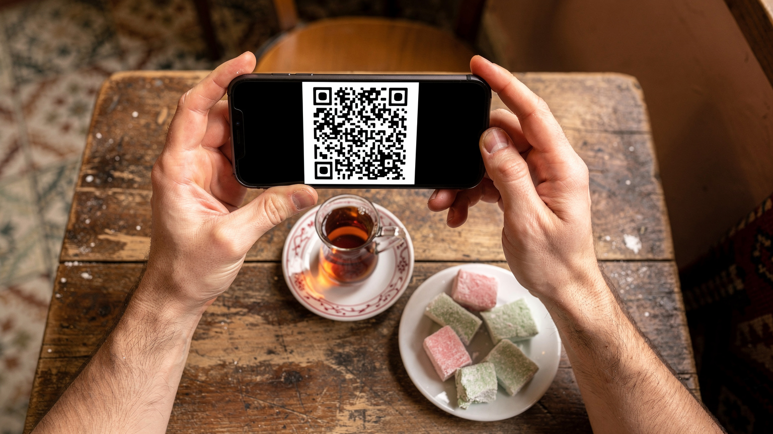 يد رجل تحمل هاتفًا ذكيًا يعرض رمز QR بينما يجلس في مقهى تركي تقليدي، مع فنجان من الشاي التركي والحلويات على الطاولة الخشبية أمامه.