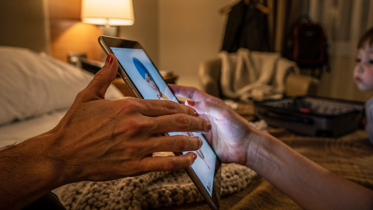 A close-up of a parent's hands carefully downloading a movie onto a child's tablet while sitting in a cozy hotel room, late at night, in preparation for the next day's travel.