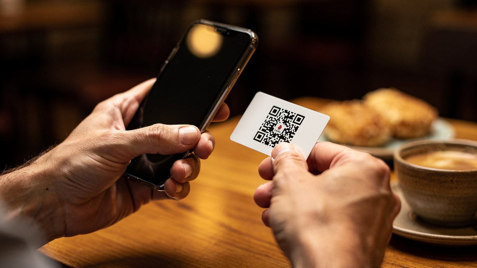 Manos de una persona sosteniendo un smartphone, acercándolo a un código QR impreso en una tarjeta, en un café de Asunción, ilustrando el proceso de activación de una eSIM.