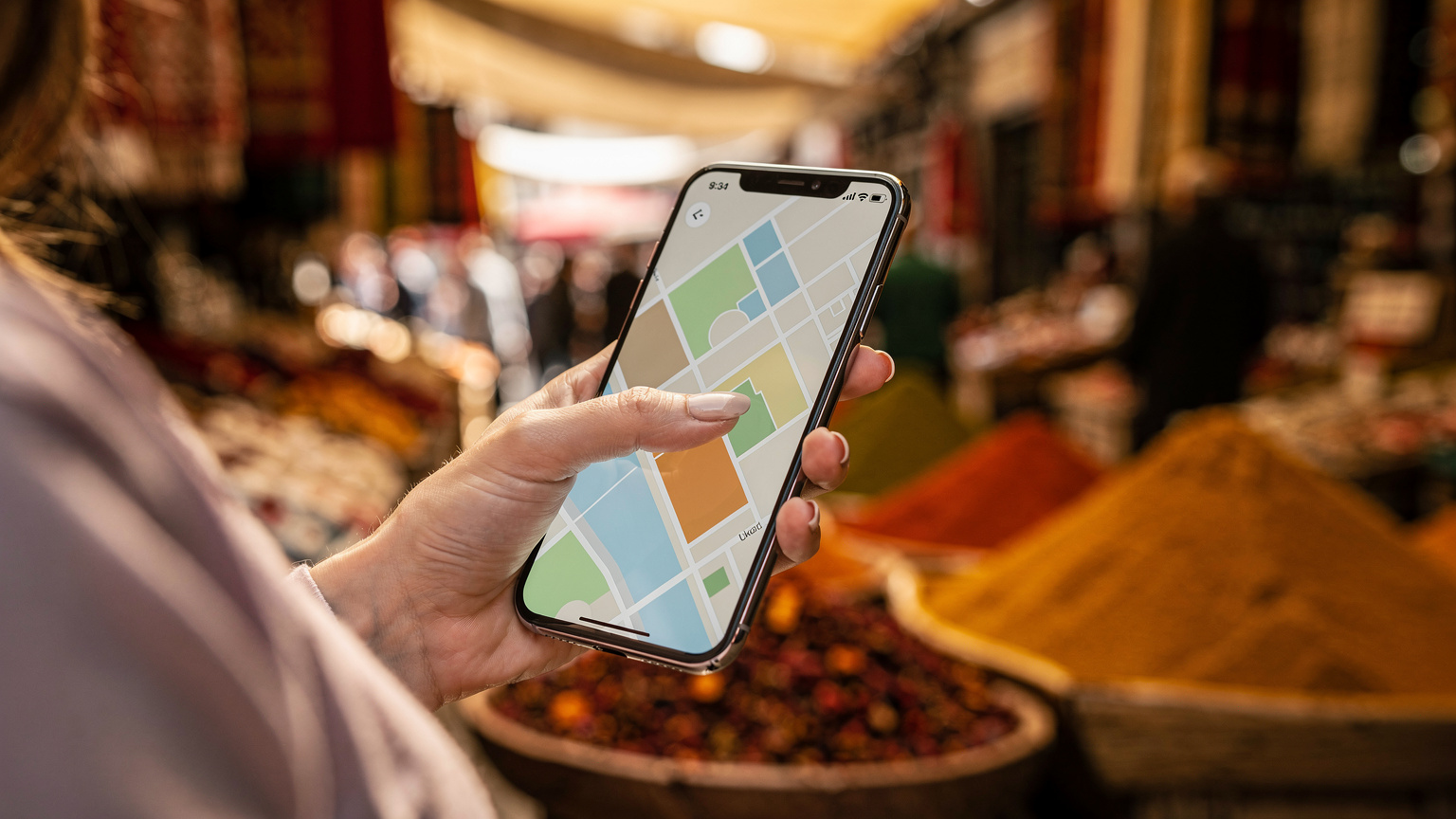 Mână apropiată a unei femei care navighează pe o aplicație de hărți pe un smartphone, cu fundalul estompat al unui bazar tradițional turcesc plin de culori și oameni.