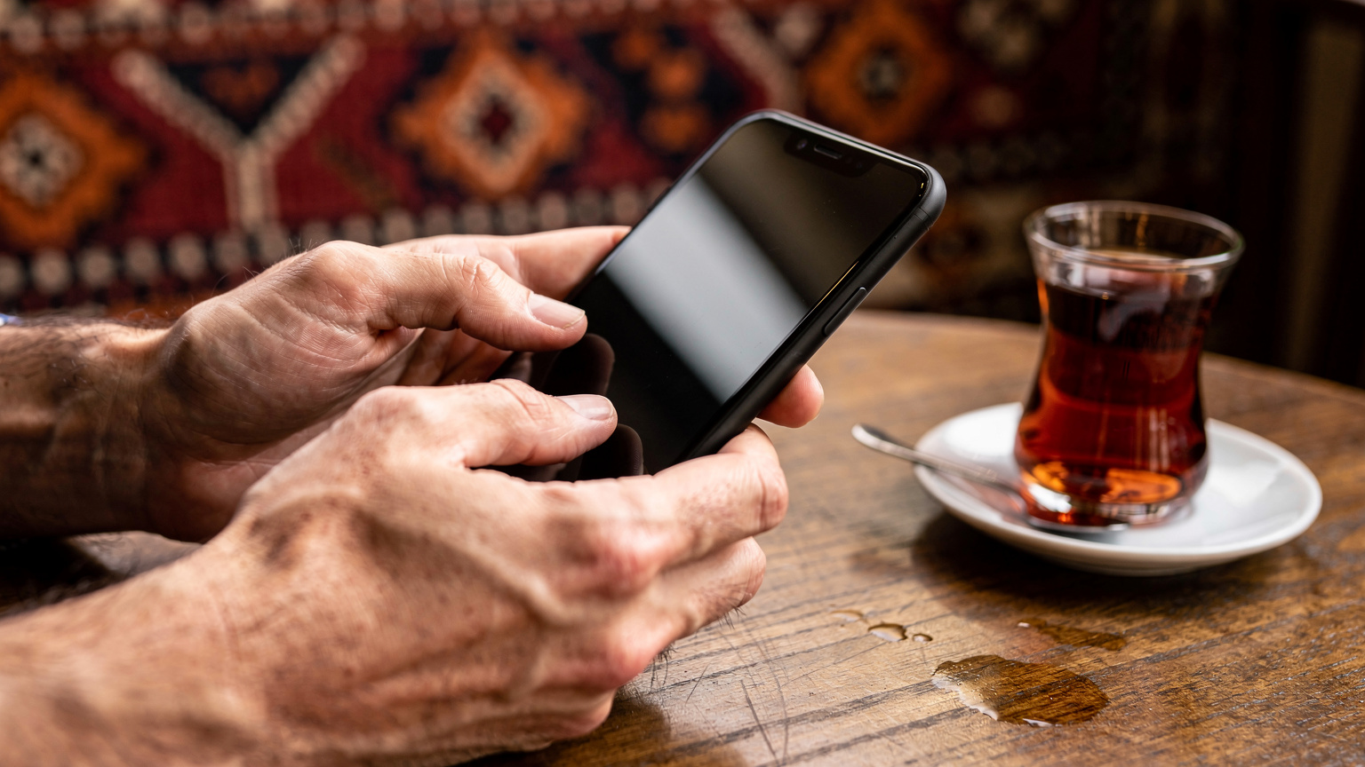 Close-up of human hands holding a smartphone, with fingers tapping on the screen. The screen is blurred to avoid showing specific UI elements, focusing on the interaction itself. The background is a slightly out-of-focus cafe interior in Istanbul, with a traditional Turkish tea glass on the table.