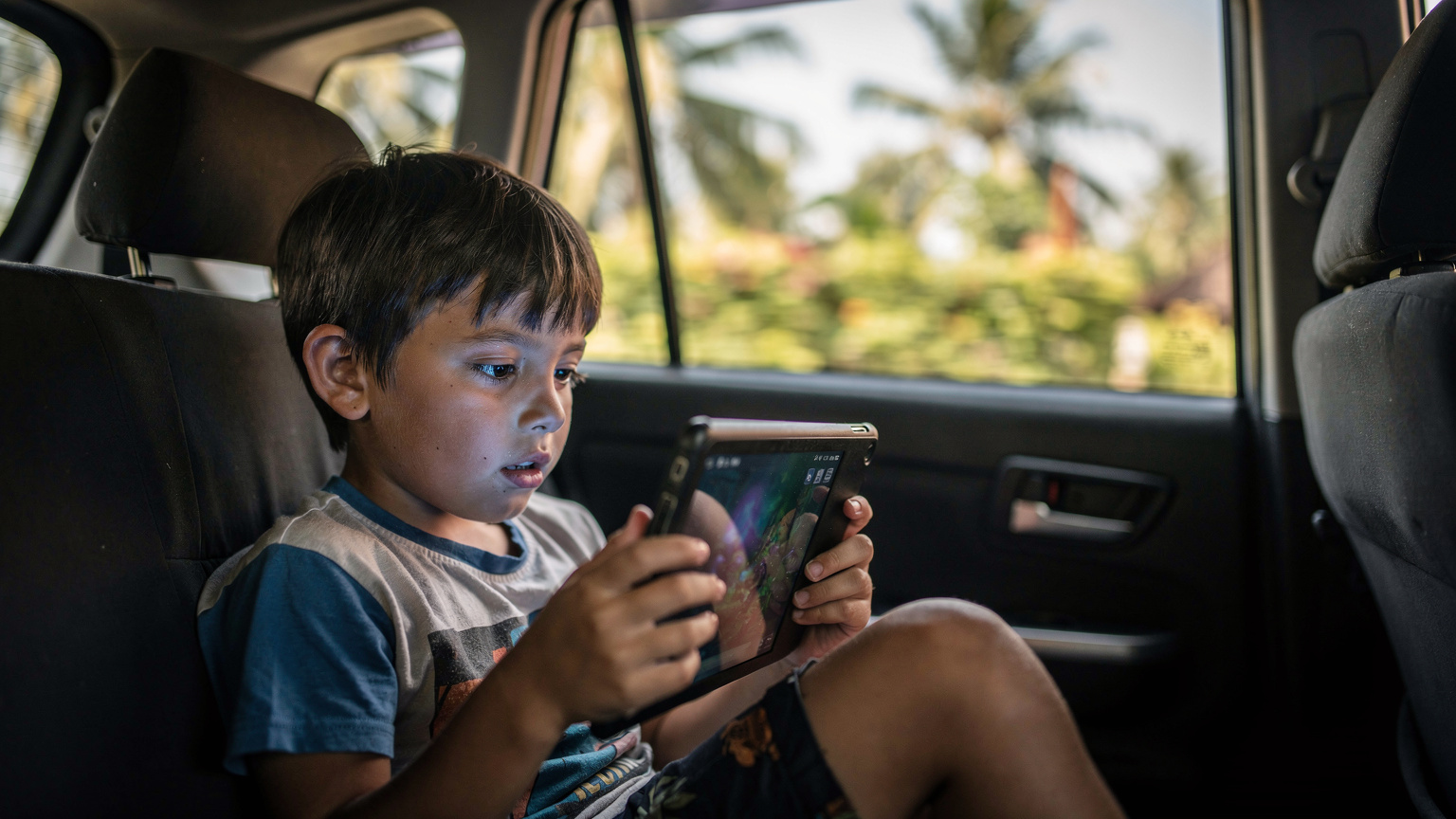 A child, around 7 years old, engrossed in a tablet game while sitting comfortably in the backseat of a moving taxi in Bali. The tablet screen is brightly lit, reflecting slightly in the child's eyes. Outside the window, blurred views of lush Balinese landscapes pass by, suggesting travel.