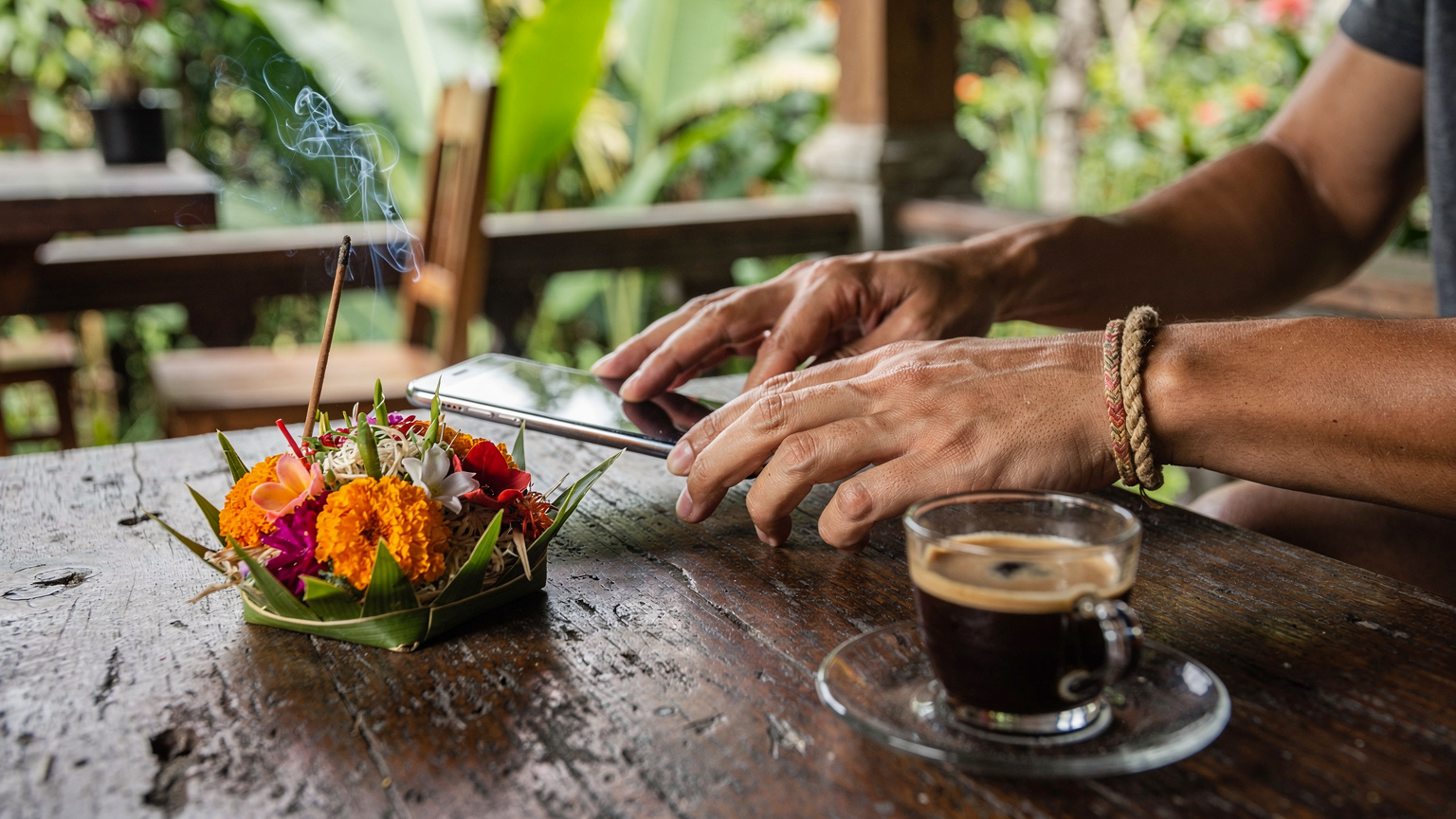 Close-up of a traveler's hands gently placing a smartphone down on a wooden table next to a Balinese offering (Canang Sari) and a small glass of kopi luwak, signifying successful eSIM activation and a moment of cultural immersion.