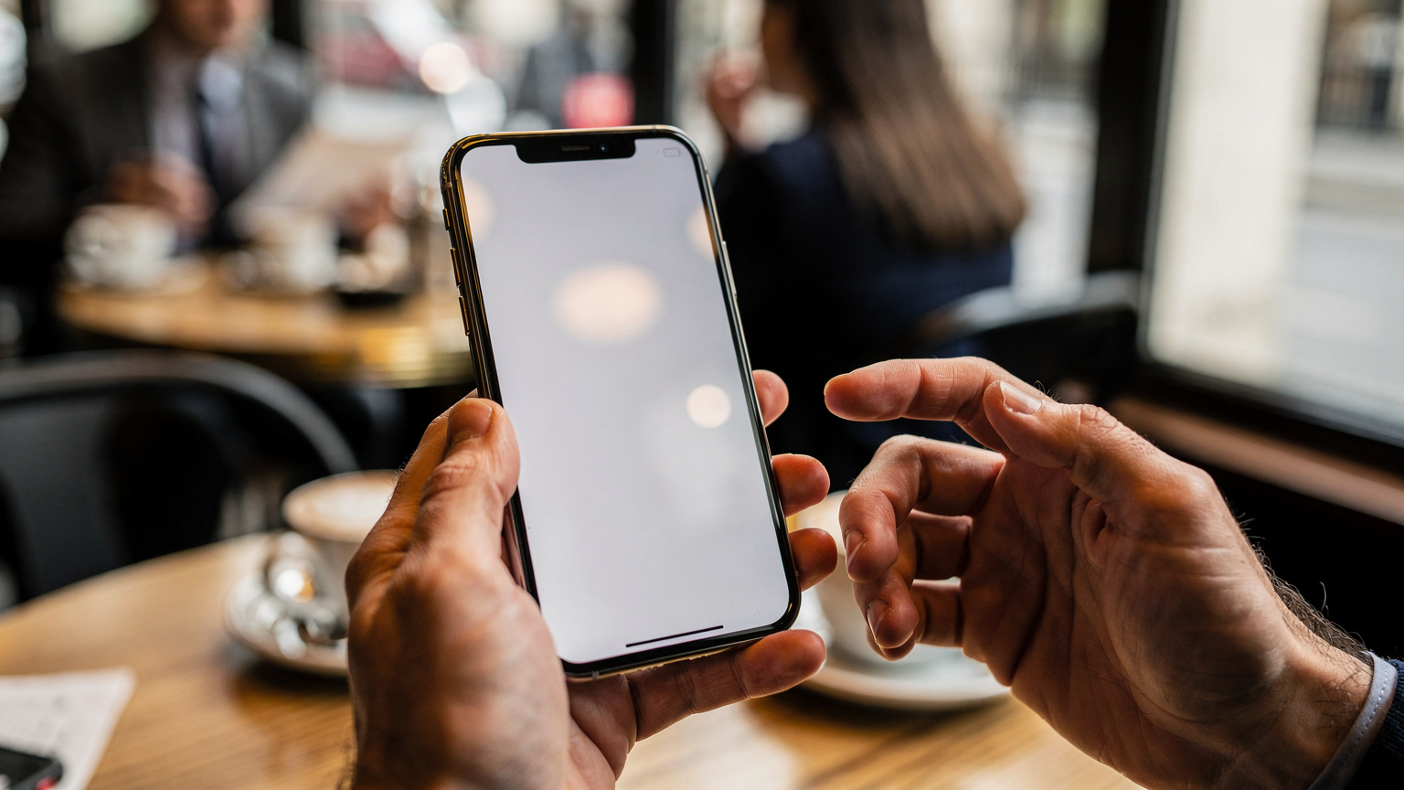Un écran de smartphone affichant les paramètres de la carte SIM, avec l'option "Ajouter un forfait cellulaire" visible, tenu par des mains humaines dans un café animé du quartier de Marais à Paris.