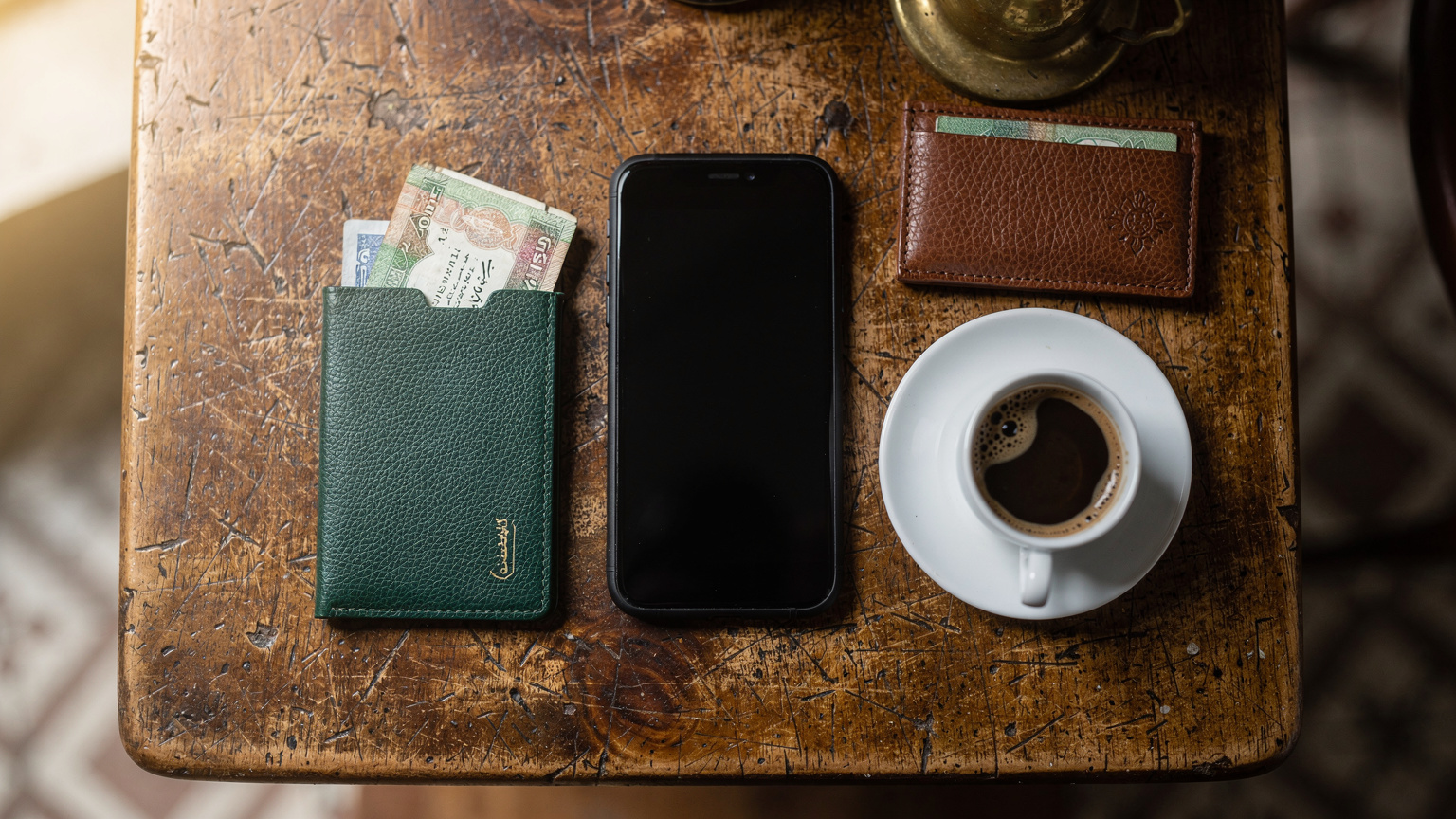 عرض علوي (Flat lay) يظهر هاتفاً ذكياً، جواز سفر عراقياً، محفظة صغيرة مفتوحة تكشف عن عملات الدينار العراقي، وفنجان قهوة عربية تقليدية، كلها موضوعة على طاولة خشبية عتيقة في مقهى ببغداد.