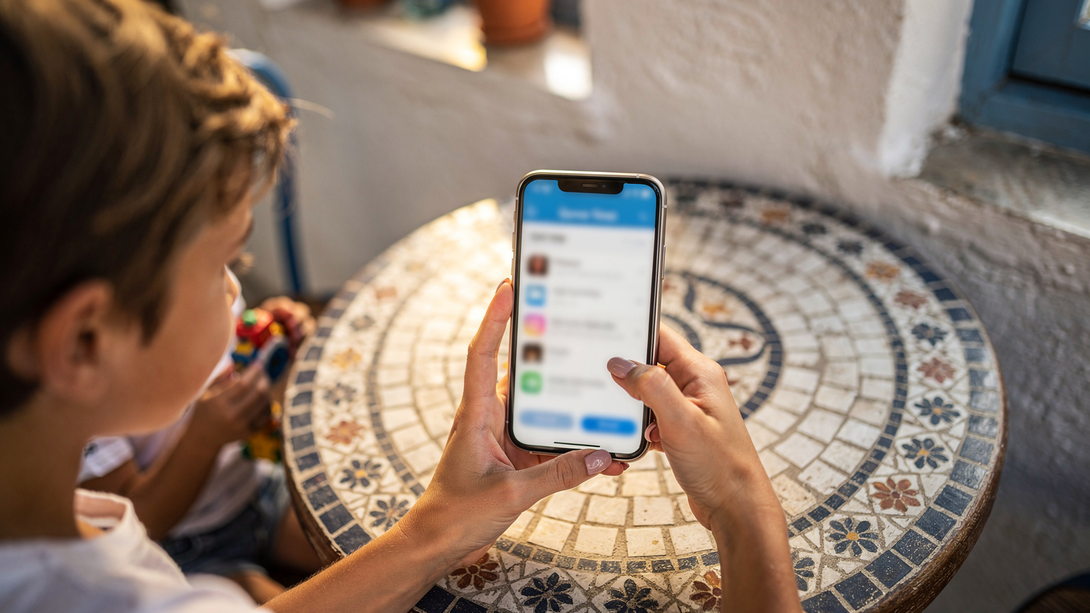 يد أم تمسك بهاتف ذكي يعرض إعدادات وقت الشاشة، بينما يجلس طفلها الصغير بجانبها في مقهى يوناني تقليدي في منطقة بلاكا بأثينا.