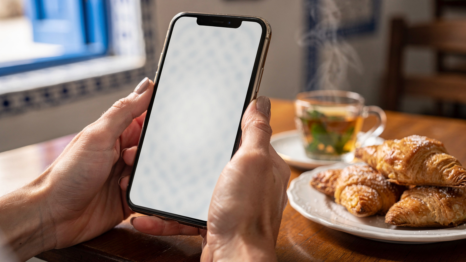 Un plan rapproché d'un smartphone affichant un écran de confirmation d'activation d'eSIM, tenu par des mains féminines avec un vernis à ongles discret. L'arrière-plan est légèrement flou, montrant une tasse de thé à la menthe et des pâtisseries tunisiennes sur une table en bois d'un café traditionnel à Sidi Bou Saïd. La lumière est douce et naturelle, filtrant à travers une fenêtre. L'objectif est un 85mm, avec une très faible profondeur de champ f/1.4, pour isoler le téléphone et les mains. Le style est une photographie éditoriale de type 'lifestyle' de voyage. Aucune interface utilisateur visible, aucun logo lisible.