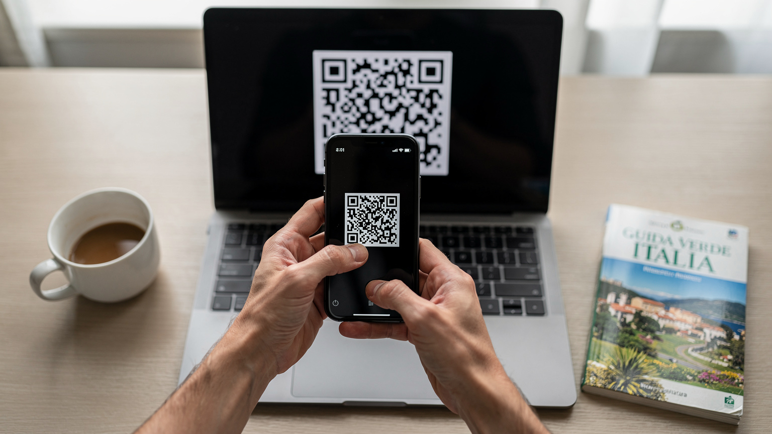 Primo piano delle mani di una persona che tiene uno smartphone e scansiona un codice QR visualizzato su un portatile sottile, con una tazza di caffè e una guida turistica dell'Italia sullo sfondo sfocato, in un ambiente domestico o di hotel.