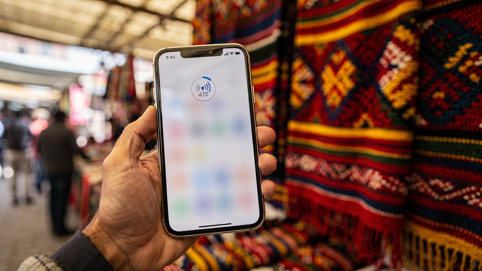A close-up shot of a smartphone displaying a network signal indicator, held by a person in a local market in La Paz, Bolivia, with colourful textiles in the background.
