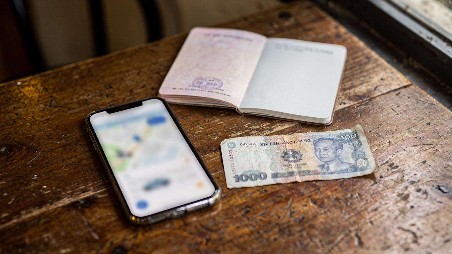 An overhead shot of a phone displaying a ride-sharing app, a passport, and a local currency note on a wooden cafe table in Ho Chi Minh City, Vietnam. Natural light from a window.