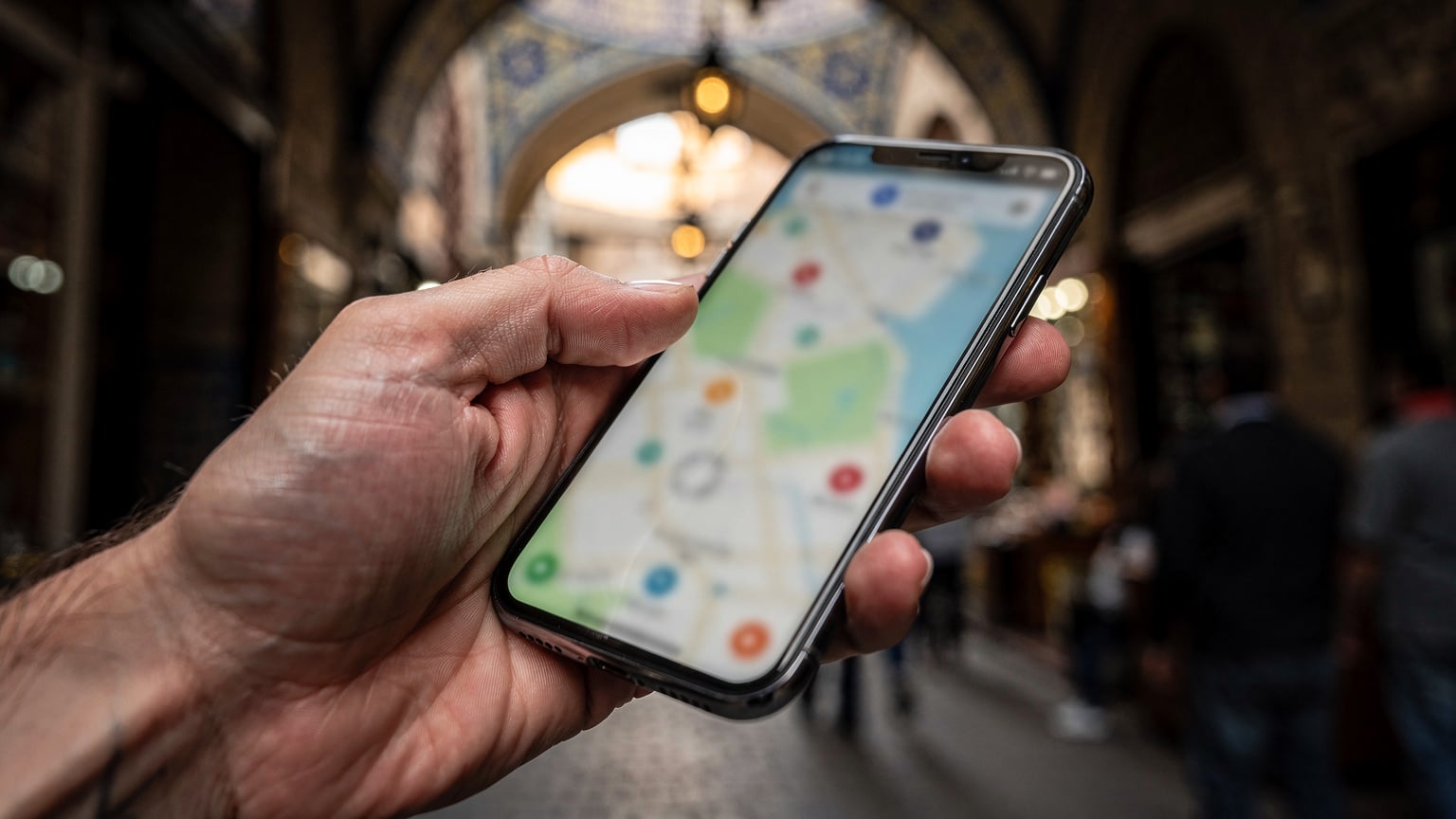 A close-up of a traveller's hand holding a smartphone, looking at a map application on a crowded street near the Grand Bazaar in Istanbul. The screen is visible but blurred, showing only a map interface.