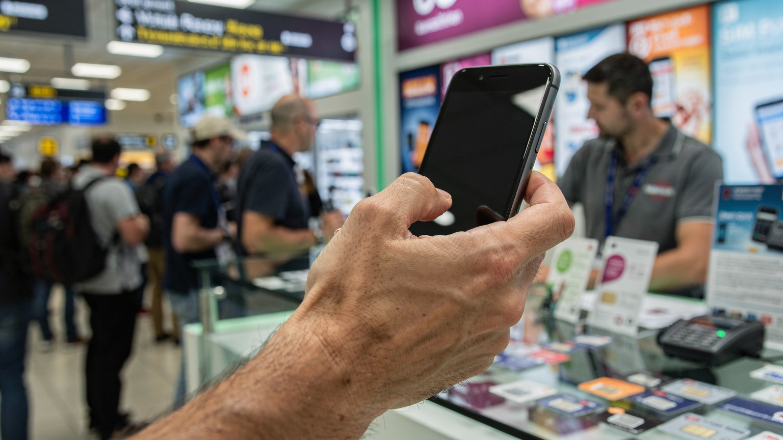 Mână masculină ținând un telefon Android, privind cu frustrare ecranul, într-un birou de vânzări SIM aglomerat dintr-un aeroport.