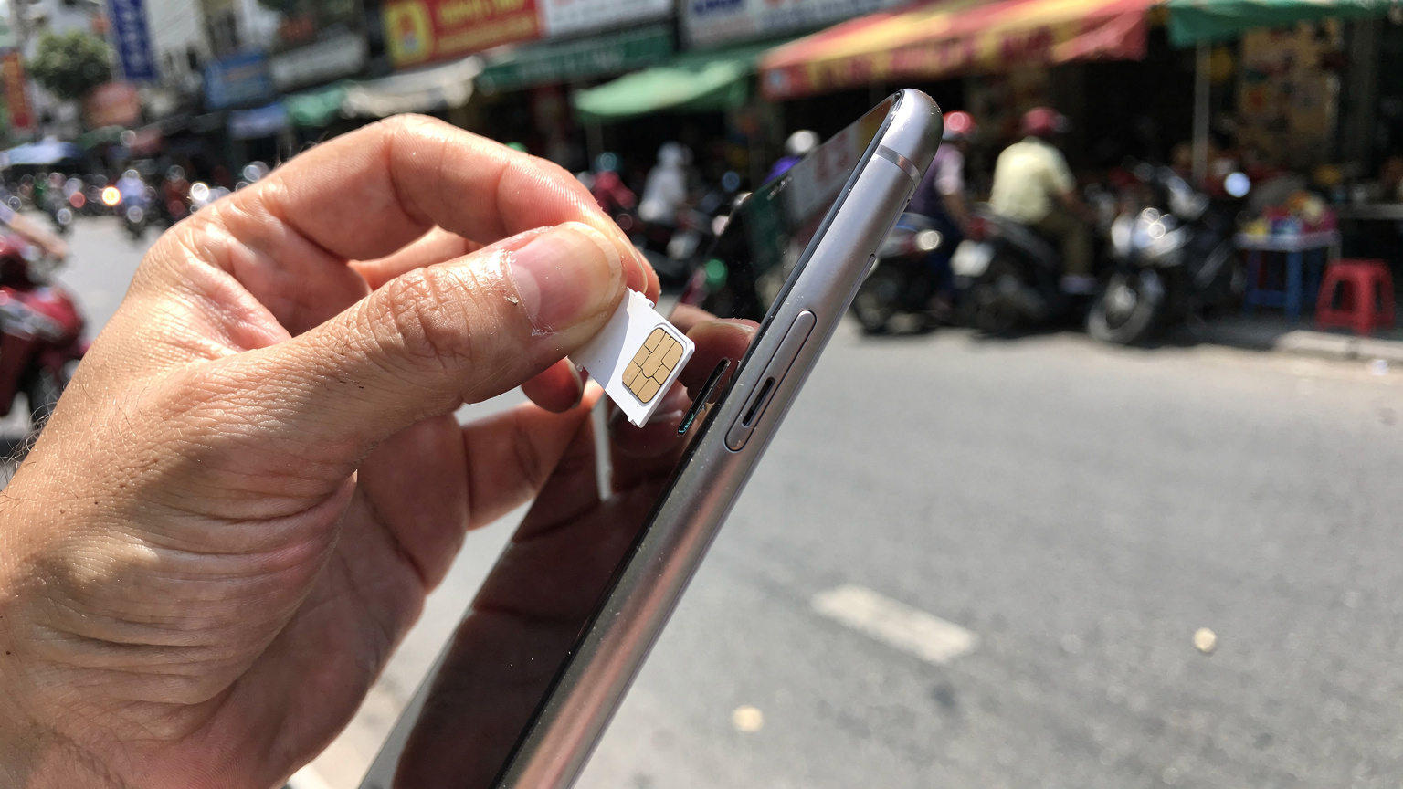 Eine Nahaufnahme einer Hand, die eine lokale SIM-Karte in einen Smartphone-Slot einlegt, mit einem verschwommenen Hintergrund einer belebten Straße in Südostasien.