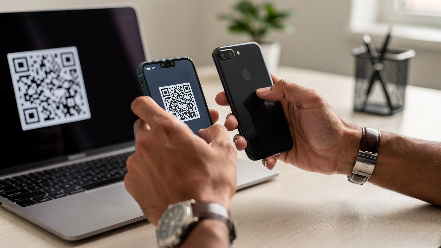 لقطة مقربة لأيدي شخص عربي يرتدي ساعة يد أنيقة وهو يمسح رمز QR ضوئيًا من شاشة كمبيوتر محمول باستخدام هاتف ذكي، مع خلفية مكتبية عصرية مضاءة بشكل ناعم.