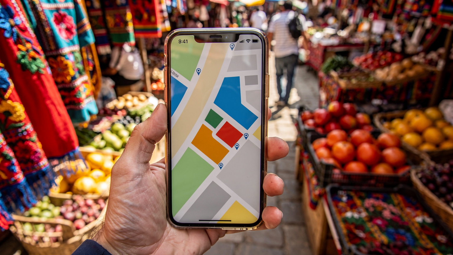 Un teléfono móvil mostrando una aplicación de mapa en el mercado de Chichicastenango, Guatemala, con telas coloridas y productos locales al fondo.